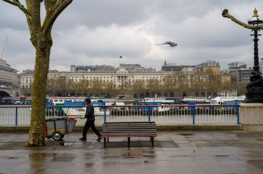 LONDON - 21 Nisan 2023: Gri bir günde arka planda Thames nehri bulunan ve şehrin herkes için temiz ve güzel kalmasını sağlayan Southbank boyunca bir araba iten bir sokak temizleyicisinin ithafına tanık olun..