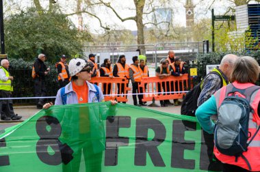LONDON - 22 Nisan 2023: Cesur bir Soykırım İsyanı protestocusu yolun karşısında bir pankart tutarken, yakındaki bir şantiyeden, yüksek ceketli adamlar Londra 'daki sahneyi izliyorlar..