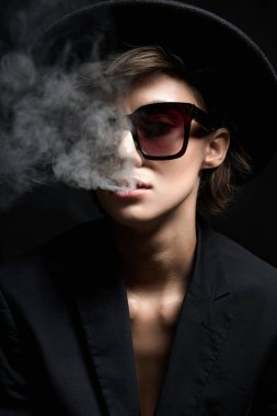 Güneş gözlüklü ve şapkalı güzel bir kadın. Takım elbiseli güzel bayan ağzından duman salıyor.