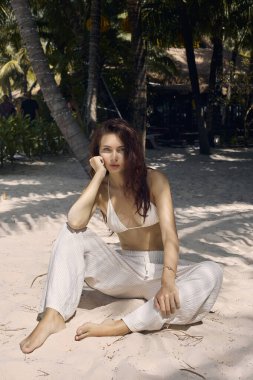 Güzel kız palmiyelerin altındaki kumda oturuyor. Kumsalda Bikini 'deki seksi esmer kadın.