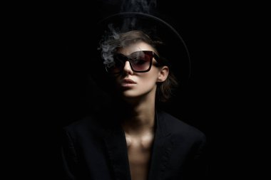 Güneş gözlüklü ve şapkalı güzel bir kadın. Takım elbiseli güzel bayan ağzından duman salıyor.