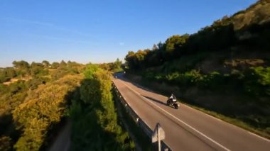 Dynamic fpv drone shot chasing Motorcycle driving under the bridge while sunset on serpentine road during golden hour, driver with white long sleeve shirt . followed with high speed fpv drone, 4k