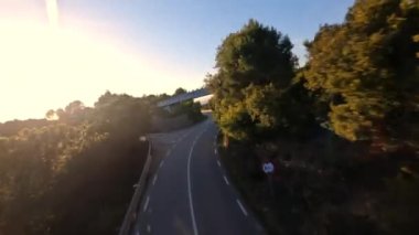 Dynamic fpv drone shot chasing Motorcycle driving under the bridge while sunset on serpentine road during golden hour, driver with white long sleeve shirt . followed with high speed fpv drone, 4k
