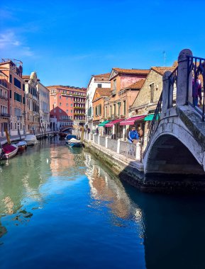 Venedik, İtalya - Nisan 2023. Yelkenli Venedik kanalları ve güneşli bir günde sokaklarda yürüyen turistler.