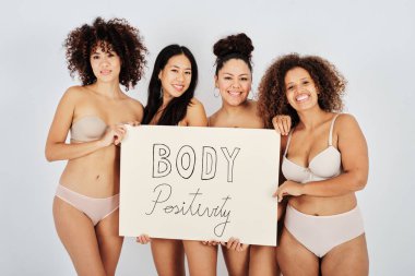 İç çamaşırlı mutlu kadın modeller gülümseyerek kameraya bakıyor ve gri arka planda vücut pozitifliği yazısını gösteriyor.