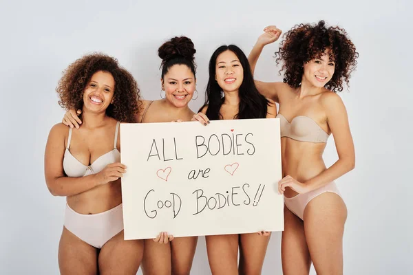 stock image Group of cheerful diverse girlfriends in underwear looking at camera with smile and showing All Bodies Are Good Bodies poster against gray background