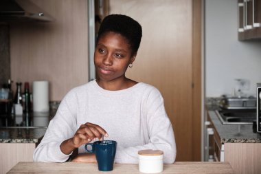 Çağdaş mutfakta masada dinlenen düşünceli siyah kadın kahvaltıda bir fincan sıcak aromatik içki içerken.