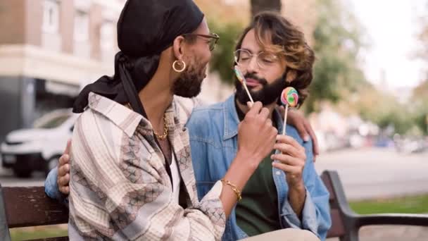 路上でベンチに座って笑っている間にロリポップを食べる多人種間の同性愛者のカップルのスローモーションビデオ — ストック動画