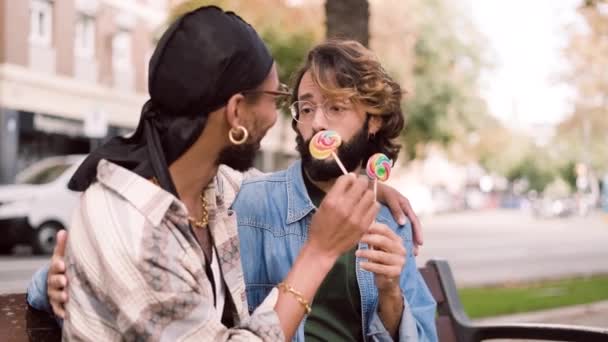 一对快乐的多文化同性恋夫妇一边吃棒棒糖一边坐在街上的长椅上聊天的慢镜头 — 图库视频影像