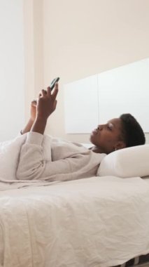 Yatakta yatarken gülümseyen Afrikalı bir kadının cep telefonunu kullanmasının dikey yavaş çekim videosu.