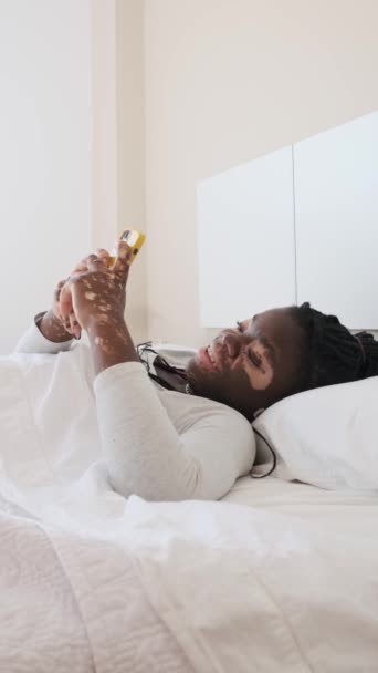 用手机从床上拍摄的一个快乐而懒惰的患白癜风的非洲女人的慢动作垂直录像 — 图库视频影像