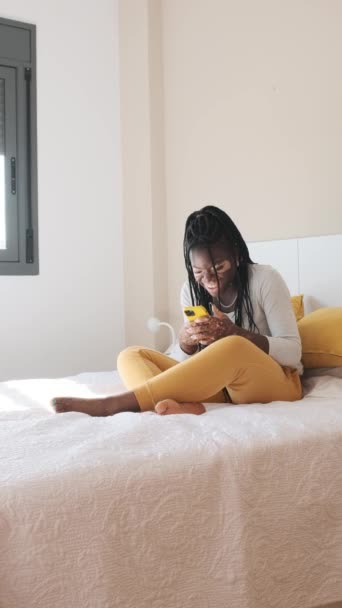 一个笑着和放松的非洲妇女的慢动作垂直视频 她的皮肤患有白粉风 她用手机坐在床上 — 图库视频影像