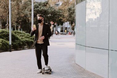 Sokakta cep telefonunu kullanırken yüz koruyucu maskeli beyaz bir adam patenle kayıyor.