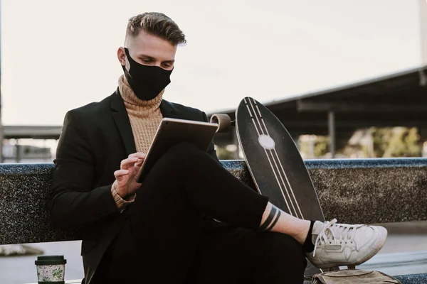 Man with facial mask using a tablet sitting in an urban bench next to a skateboard