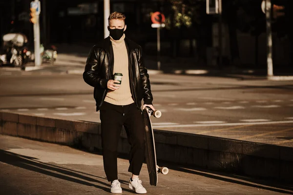 Portrait of a stylish man with a facial mask holding a take away coffee standing with a skateboard