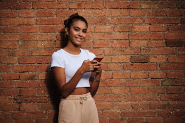Kadın kameraya bakıyor ve dışarıdaki bir cep telefonunu tuğla duvara karşı kullanırken gülümsüyor. Teknoloji ve sosyal medya kavramı.