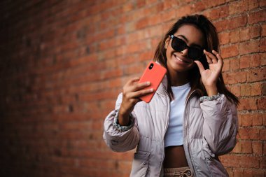 Güneş gözlüklü bir kadın cep telefonuyla duvara selfie çekerken gülümsüyor. Teknoloji ve sosyal medya kavramı.