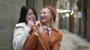 Şehirdeki dar bir sokakta kalp şeklinde lolipop yerken Asyalı bir lezbiyenin kucaklaşmasının yavaş çekim videosu.