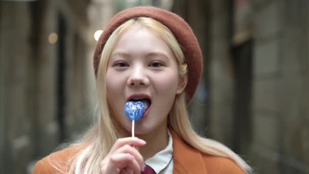 一个时髦的金发亚洲女人在街上吃蓝色心形棒棒糖的慢镜头 — 图库视频影像