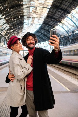 Tren istasyonunda selfie çeken mutlu bir çiftin dikey portresi.