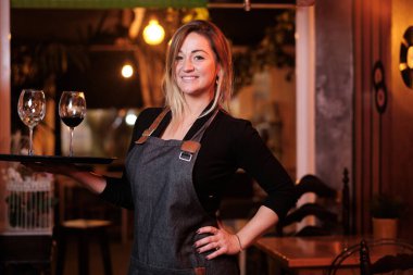 Garson, restoranda şarap kadehiyle tepsiyi tutarken kameraya bakıp gülümsüyor..