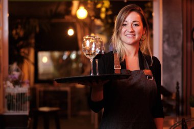 Kadın garson, şarap kadehiyle tepsiyi tutarken kameraya bakıp gülümsüyor..