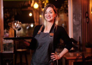 Üniformalı garson, elinde şarap kadehiyle bir restoranda beklerken kameraya gülümsüyor..