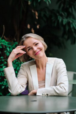 Bir kadın dışarıda bir masada otururken kameraya bakıyor ve gülümsüyor..