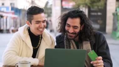 Slow motion video of a happy gay couple buying online using a laptop while sitting in a cafeteria