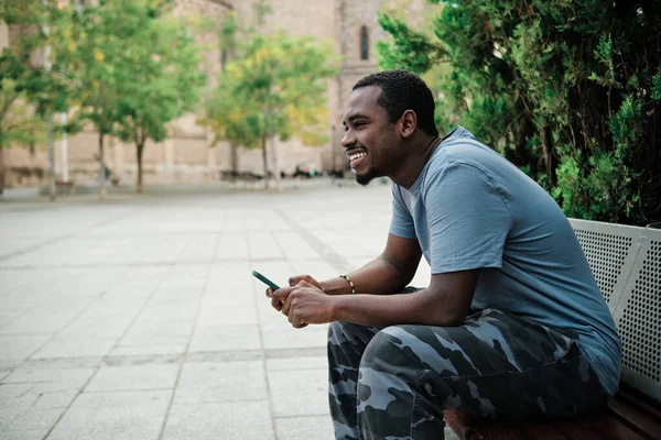 Man smiling while using mobile phone sitting on a bench outdoors. Technology concept.