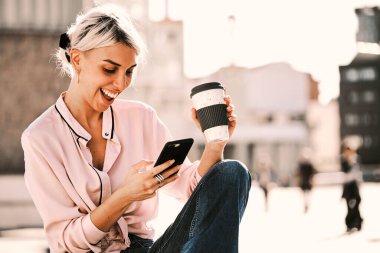 Woman laughing while using a mobile phone and drinking coffee outdoors on the street. Technology concept.