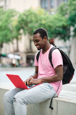 Man with backpack smiling while using a laptop sitting on a bench outdoors. Technology concept.