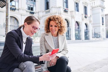 Two women using mobile phone together while sitting on a bench on the street. Technology concept.