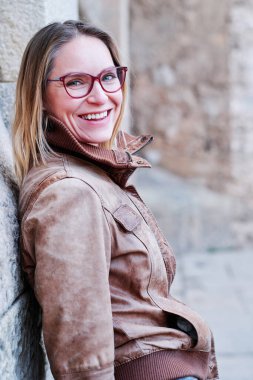 Woman with eyeglasses looking at camera and smiling while posing leaning on a wall outdoors.