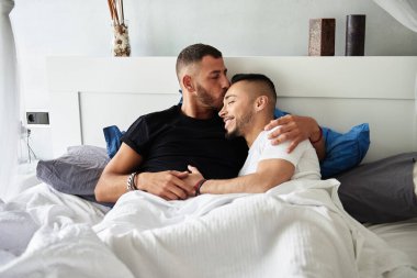 Male gay couple smiling and cuddling while relaxing lying in bed together at home. Lgbt concept.
