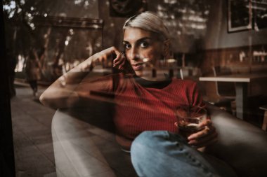 View from outside of a woman looking at camera while enjoying drinking beverage sitting in a restaurant.