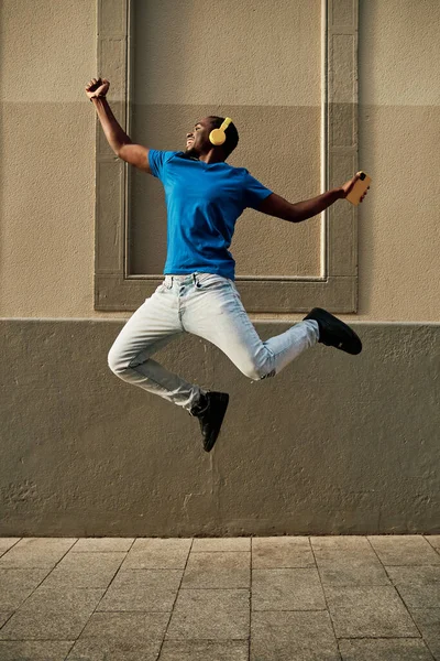 Excited man jumping while enjoying listening music with headphones and mobile phone outdoors. Technology concept.