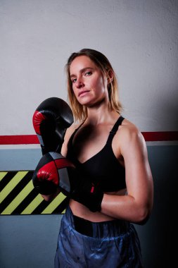 Sporty woman looking at camera while posing with boxing gloves indoors. Sports concept.
