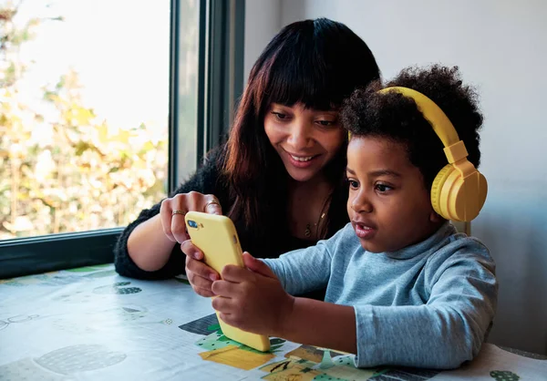 Domestic scene of a young latin mother and son using a mobile and headphones sitting at home