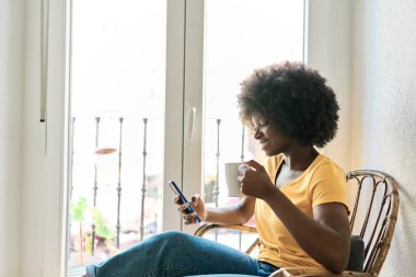 Afrikalı bir kadın evdeki tahta sandalyede rahat rahat otururken kahve içiyor.