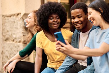 Caddede oturan cep telefonunu kullanan mutlu çok ırklı arkadaş grubu