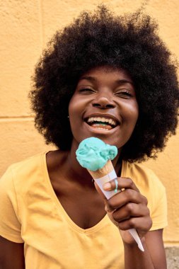 Mutlu Afrikalı kadın dışarıda dondurma yiyor. Sarı bir şehir duvarının yanında.