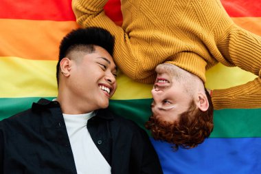 lgbt bayrağında yatarken gülümseyen çok kültürlü eşcinsel bir çiftin üst görünümü.