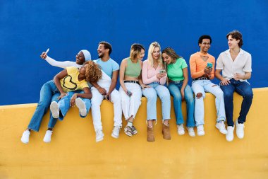 Renkli bir duvarda oturan çok ırklı gençlerin telefon kullanışının ön yüzü.