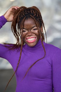 Mor bluzlu neşeli bir kadın, güneşli bir günün tadını çıkarıyor, örgülü saçlarına neşeyle dokunuyor.