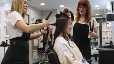 Kuaförlerden oluşan bir ekibin, kuaförde oturan Çinli bir kadının saçlarını kıvırmasını gösteren yavaş çekim videosu.