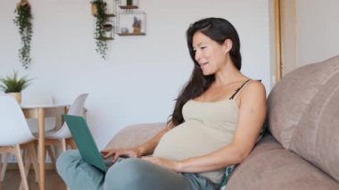 Asyalı hamile bir kadının evdeki bilgisayarla video görüşmesi yaparken yavaş çekim videosu.