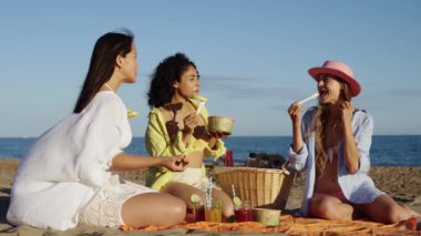 Three women are sitting on the beach, eating and drinking. They are wearing hats and are smiling