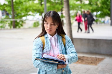 A chinese student wearing a blue jacket and white shirt is looking at her phone while holding a stack of books
