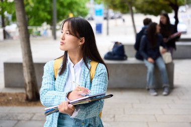 chinese student wearing a blue jacket and white shirt is holding a stack of books. She looks thoughtful and is standing in front of a bench